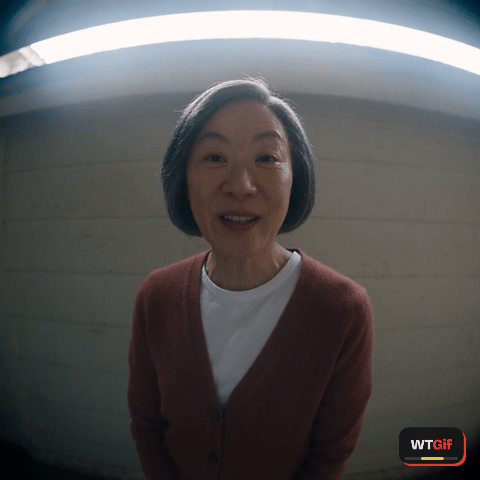 On a wide doorbell camera, a woman in her sixties with a short grey bob and a burgundy cardigan stops talking mid-word, her polite smile drops, she tilts her head a little with her eyebrows up, then lowers her chin and looks flatly down into the lens.