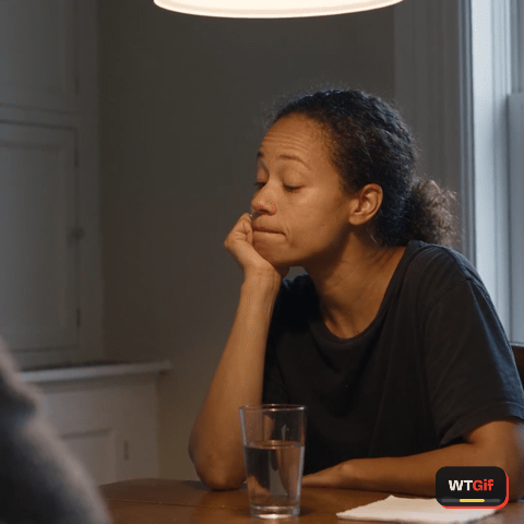 A phone drifts across a dinner table to a woman in her thirties with curly hair tied back, chin on her hand, who blinks, lifts one eyebrow, tilts her head, and slides her eyes to the camera, unimpressed.