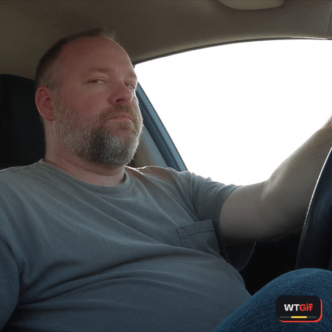 Filmed from the passenger seat of a car stopped at a light, a heavyset man in his fifties with a short grey beard blinks once, his eyes slide toward the phone, then his head follows in a slow turn with his eyebrows raised, and he holds the look.