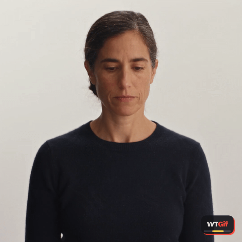 A woman in her forties with a low bun and a black sweater looks down, slowly widens her eyes, then lifts them to the camera with a small open mouth and a hand flat on her chest, and gives one slow deadpan head shake.