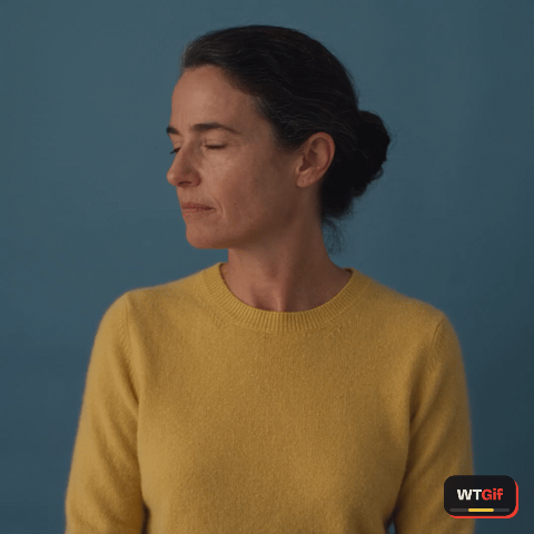 A woman in her mid forties with black hair in a low bun and a little grey at the temples, in a plain yellow sweater, turns slowly to the camera against a cobalt backdrop with a tight half smile and an amused squint, then gives one tiny sure, okay nod and holds it.