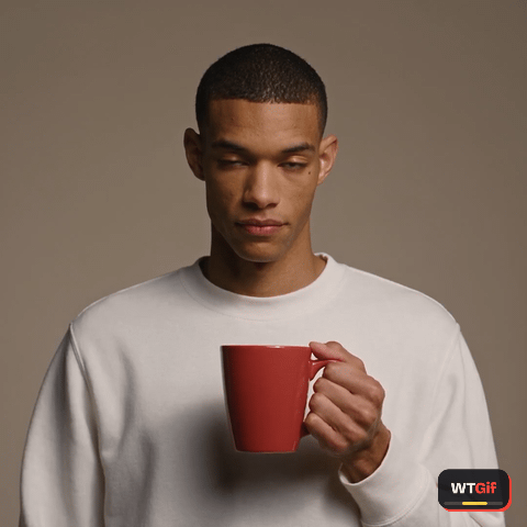 A young man with deep brown skin and a low fade, wearing a plain white sweatshirt, glances up from a red mug, looks back down, then snaps his eyes to the camera wide eyed in a double take and holds the deadpan stare.