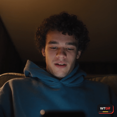 Seen from a phone propped in his lap, a young man with dark curly hair and a navy hoodie goes from a half smile at a video to wide eyes and a slack jaw as he lifts his gaze to the lens, lit only by a lamp and the screen glow.